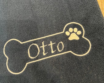 Könnte beinhalten: Ein schwarzes Hundebett mit einem beigefarbenen, knochenförmigen Flecken, auf dem der Name "Otto" und ein Pfotenabdruck gestickt sind.
