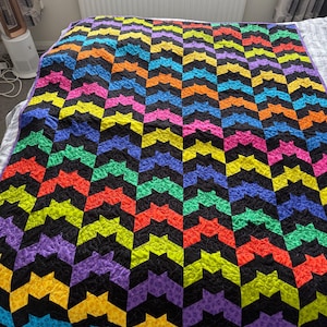 May include: A quilt featuring a repeating chevron pattern made of vibrant neon-coloured fabric pieces in yellow, orange, red, pink, purple, blue, and green, all separated by solid black fabric segments with visible quilted stitching throughout.