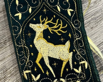 May include: A dark green, rectangular embroidered item with a gold deer design. The deer is centered within a gold-lined frame, surrounded by gold floral accents and stars. A gold ribbon is attached to the side. The item has a dark green border.
