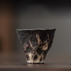 Puede incluir: Una taza de té de cerámica oscura hecha a mano con una superficie texturizada única. La taza presenta una forma acampanada y un borde irregular. El exterior tiene un patrón moteado de negro y beige, creando una estética rústica.