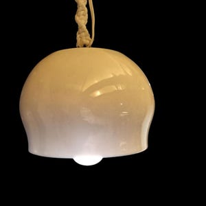 May include: A white ceramic pendant light with a white cord hanging from the ceiling. The light is turned on and illuminates the room.