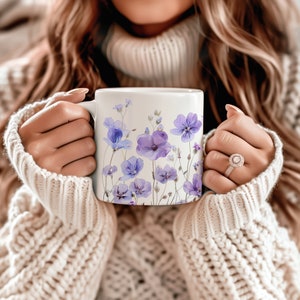 May include: A white ceramic mug with a floral design featuring purple and blue flowers. The mug is held by a person wearing a white sweater.