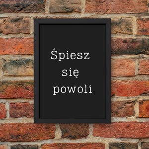 May include: A framed black sign with white text that reads "Śpiesz się powoli" against a brick wall background. The brick wall has a mix of red, brown, and orange bricks. The sign is rectangular and has a simple black frame.