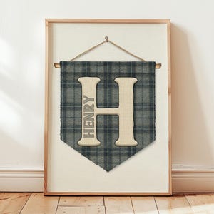 Op de afbeelding: Een ingelijste wanddecoratie met een grote letter "H" en de naam "HENRY" verticaal afgedrukt. De letter is lichtbeige van kleur, tegen een blauw-grijze geruite achtergrond. De lijst is lichtbruin.