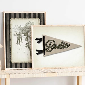 May include: Two framed prints on a wooden bench. One print is a black and white illustration of children playing. The other is a pennant with the name "Bodie" in a script font, with black ribbons.