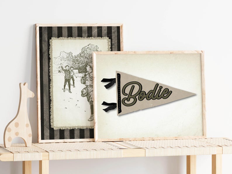 May include: Two framed prints on a wooden bench. One print is a black and white illustration of children playing. The other is a pennant with the name "Bodie" in a script font, with black ribbons.