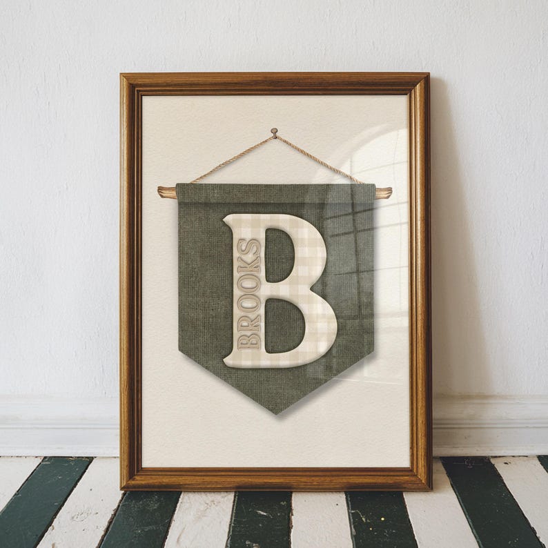 May include: Framed wall art featuring a green banner with a large, off-white letter "B" and the name "Brooks" printed vertically. The letter has a plaid pattern. The frame is a warm brown color, and the background is a neutral beige.