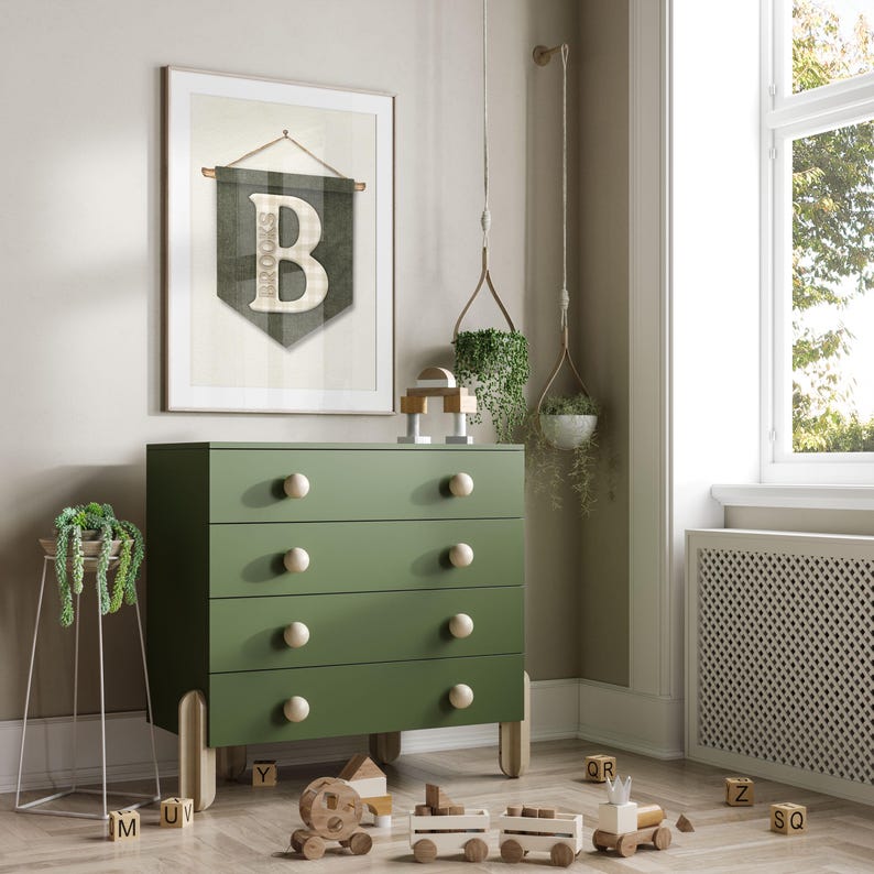 May include: A green dresser with four drawers and wooden knobs. A framed banner with the letter "B" and the name "Brooks" hangs above the dresser. Wooden toy train and blocks are on the floor. Decorative plants are also visible.