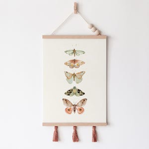 May include: A vertical wall hanging with five watercolor butterflies in shades of green, brown, and peach. The butterflies are arranged in a vertical line, with the largest butterfly at the bottom. The wall hanging is made of fabric and has a wooden dowel at the top and tassels at the bottom.