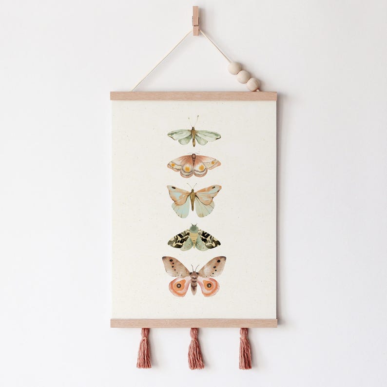 May include: A vertical wall hanging with five watercolor butterflies in shades of green, brown, and peach. The butterflies are arranged in a vertical line, with the largest butterfly at the bottom. The wall hanging is made of fabric and has a wooden dowel at the top and tassels at the bottom.