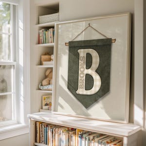 May include: Framed wall art featuring a green banner with the name "Brooks" and a large letter "B". The letter has a checkered pattern. The artwork is hanging on a wall in a room with a bookshelf and a window.