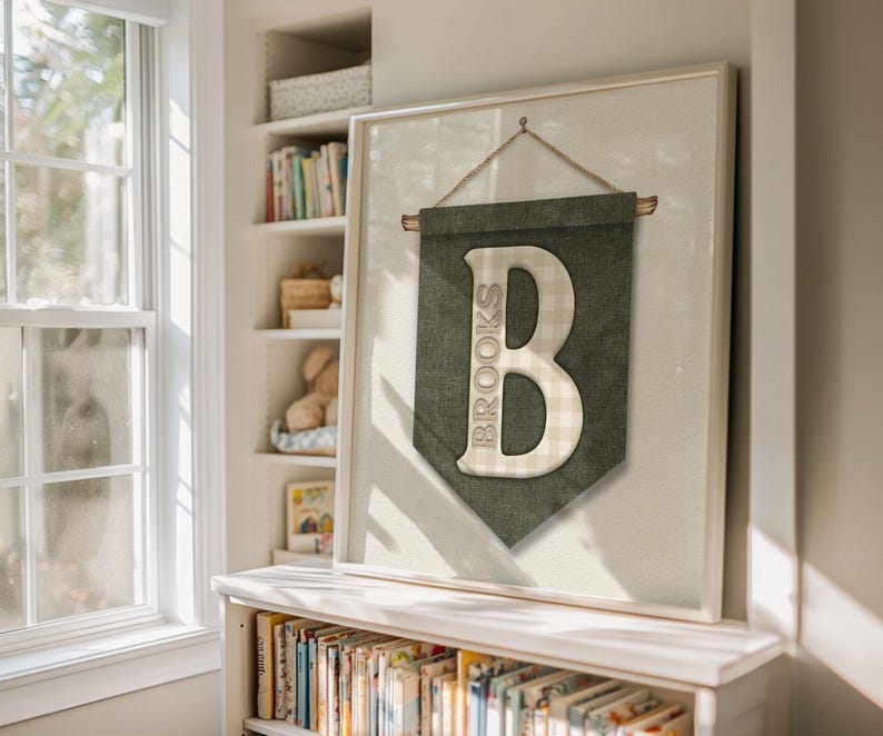 May include: Framed wall art featuring a green banner with the name "Brooks" and a large letter "B". The letter has a checkered pattern. The artwork is hanging on a wall in a room with a bookshelf and a window.