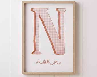 May include: A framed print with a large pink and white plaid letter N and the name "nora" written in cursive below it.