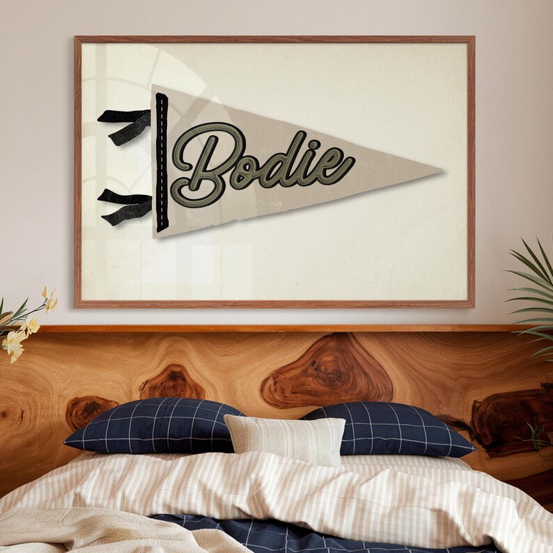 May include: A framed pennant-style wall art piece with the name "Bodie" in a script font. The pennant is a light tan color with a dark green and black outline. The frame is a medium brown color.