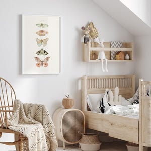 May include: A white framed print of five different butterflies with a light green, pink, and brown color scheme. The print is hanging on a white wall in a child's bedroom. The room also features a wooden bed with a white and black patterned pillow, a wicker side table, and a woven basket.