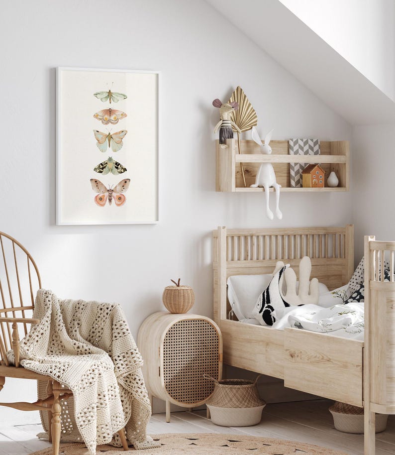 May include: A white framed print of five different butterflies with a light green, pink, and brown color scheme. The print is hanging on a white wall in a child's bedroom. The room also features a wooden bed with a white and black patterned pillow, a wicker side table, and a woven basket.
