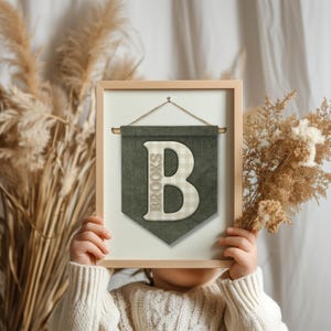 May include: A framed wall hanging with a wooden frame. The artwork features a green banner with the name "Brooks" and a large letter "B" in a plaid pattern. The banner is suspended by a rope.