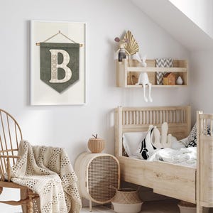 May include: A framed wall art piece with a green banner design featuring the name "Brooks" and a large letter "B". The room has a wooden bed, a woven chair, and a woven side table. A cream-colored blanket is draped over the chair.