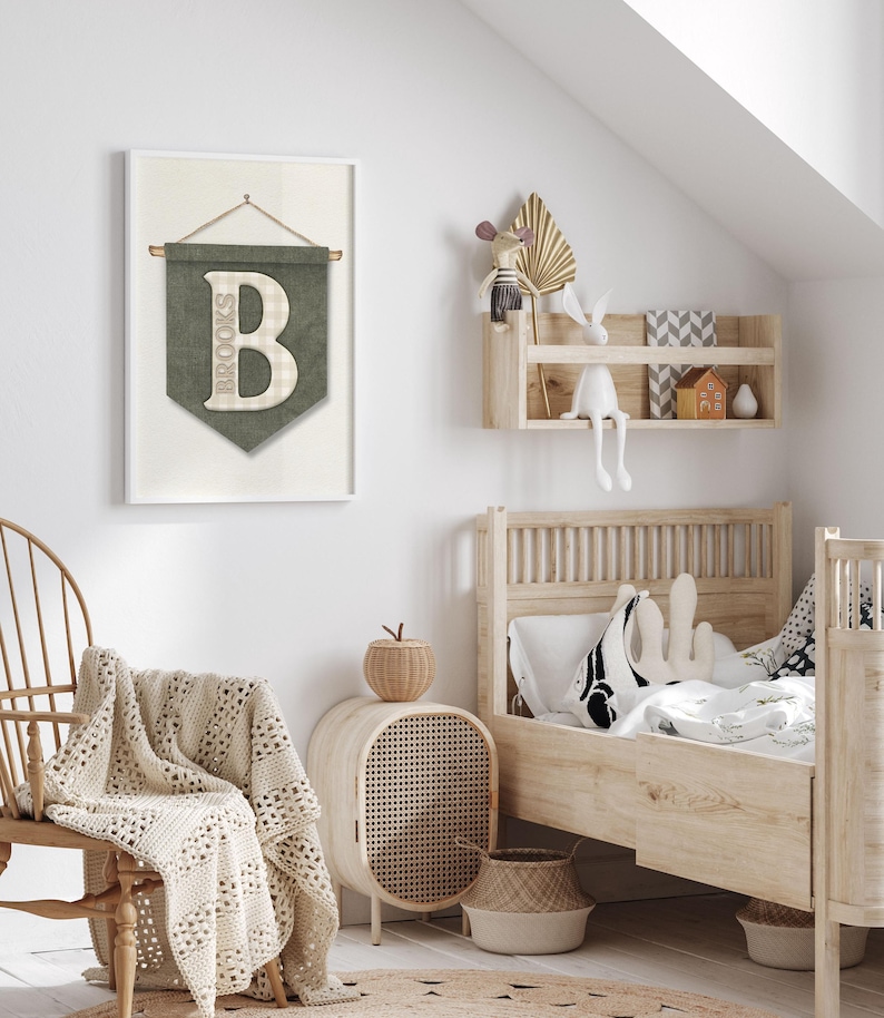 May include: A framed wall art piece with a green banner design featuring the name "Brooks" and a large letter "B". The room has a wooden bed, a woven chair, and a woven side table. A cream-colored blanket is draped over the chair.