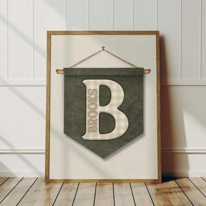 May include: A framed wall hanging featuring a green banner with the letter "B" in a beige plaid pattern. The word "BROOKS" is printed vertically on the left side of the letter. The frame is a light brown color.