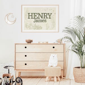 May include: A framed print with the name "HENRY James" in green lettering, displayed above a wooden chest of drawers. The room is decorated with a moon-shaped mirror, a white polar bear plush, and a potted plant. A toy car is visible on the left.