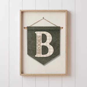 May include: A framed wall banner with a green background and a large, off-white letter "B" in the center. The letter has a plaid pattern. The word "BROOKS" is printed vertically on the left side of the letter. The banner is suspended by a wooden dowel and a rope.