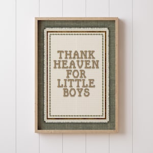 以下が含まれることがあります： 「THANK HEAVEN FOR LITTLE BOYS」の文字が刺繍された額入りのクロスステッチアート。 クリーム色の背景に茶色の糸で文字が刺繍され、緑色の布と薄茶色の木製フレームで縁取られています。 白い背景に飾られています。