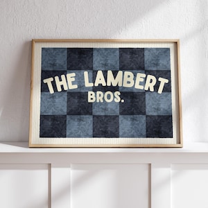 以下が含まれることがあります： 太字のオフホワイトのフォントで「THE LAMBERT BROS.」と書かれた額入りのアートワーク。背景は青とネイビーの色合いの市松模様です。フレームは明るい木の色です。