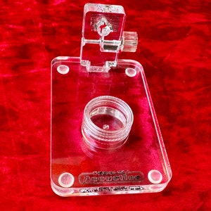 Puede incluir: Un soporte de acrílico transparente con un pequeño frasco transparente en el centro. El soporte tiene un botón de ajuste en forma de tornillo en la parte superior y las palabras "Jomolik Acoustics" impresas en la parte inferior.