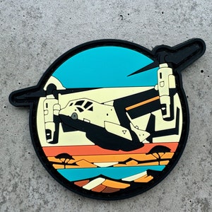 May include: A circular patch featuring a black and cream-colored aircraft design set against a backdrop of a sunset over a savanna landscape. The patch has a black border and includes shades of orange, blue, and brown.