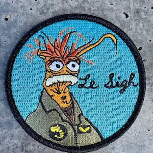 May include: Embroidered patch featuring a cartoon shrimp character with orange antennae and a white mustache, wearing a green uniform. The patch has a blue background with the words "Le Sigh" in cursive.