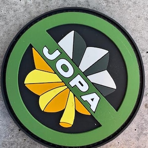 May include: A round patch with a green border and a black center. The patch features a stylized floral design in yellow, orange, white, and gray, crossed by a green bar with the white letters "JOPA".