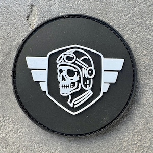 May include: A round, black patch with a white graphic of a skull wearing a pilot's helmet and goggles, set within a shield shape. The shield is flanked by stylized wings. The patch has a stitched border.