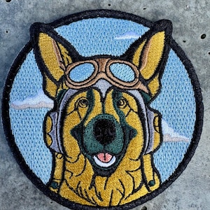 May include: Embroidered patch featuring a German Shepherd dog wearing aviator goggles and a headset. The dog is smiling with its tongue out, set against a light blue sky with white clouds. The patch has a black border.
