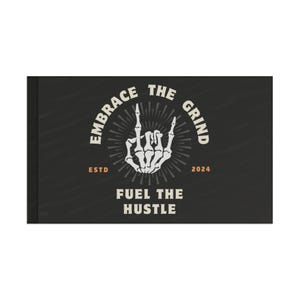 Peut inclure: Cahier noir avec le texte "EMBRACE THE GRIND" et "FUEL THE HUSTLE" en blanc. Une main de squelette blanche fait un geste de rock and roll. Le cahier comprend également le texte "ESTD 2024".