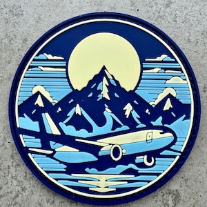 May include: A round patch featuring a mountain range, a full moon, and an airplane. The design uses shades of blue, light blue, and cream. The patch has a dark blue border and is set against a gray background.