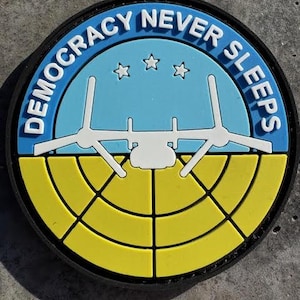 May include: A round patch with the text "DEMOCRACY NEVER SLEEPS" in white letters on a blue background. The patch features a stylized airplane and three white stars above a yellow and black segmented design.