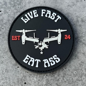May include: A round, black patch with white text and a white graphic of a military aircraft. The text reads "LIVE FAST EAT ASS" with "EST 24" in red. The patch is on a gray, textured surface.
