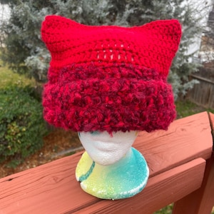May include: A handmade red cat ear hat. The hat is crocheted with a textured, fuzzy yarn in a deep red color for the brim and a smooth, bright red for the ears and top. The hat is displayed on a mannequin head.