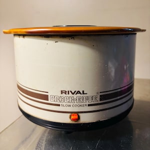 May include: Cream-colored Rival Crock-ette slow cooker with brown stripes and a brown lid. The slow cooker has the brand name and "SLOW COOKER" printed on the front. An orange light is illuminated on the front.