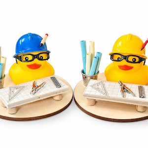 May include: Two yellow rubber ducks wearing hard hats and black glasses, each positioned on a round wooden base. They sit behind miniature drafting tables with a metal thimble holding blue and white paper rolls, a compass, and a ruler.