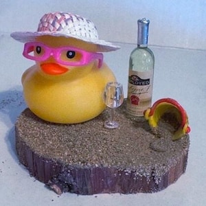 May include: A yellow rubber duck wearing a straw hat and pink glasses, next to a wine bottle and a small wine glass. The duck is on a wooden base with sand, and a small red and yellow beach bucket.