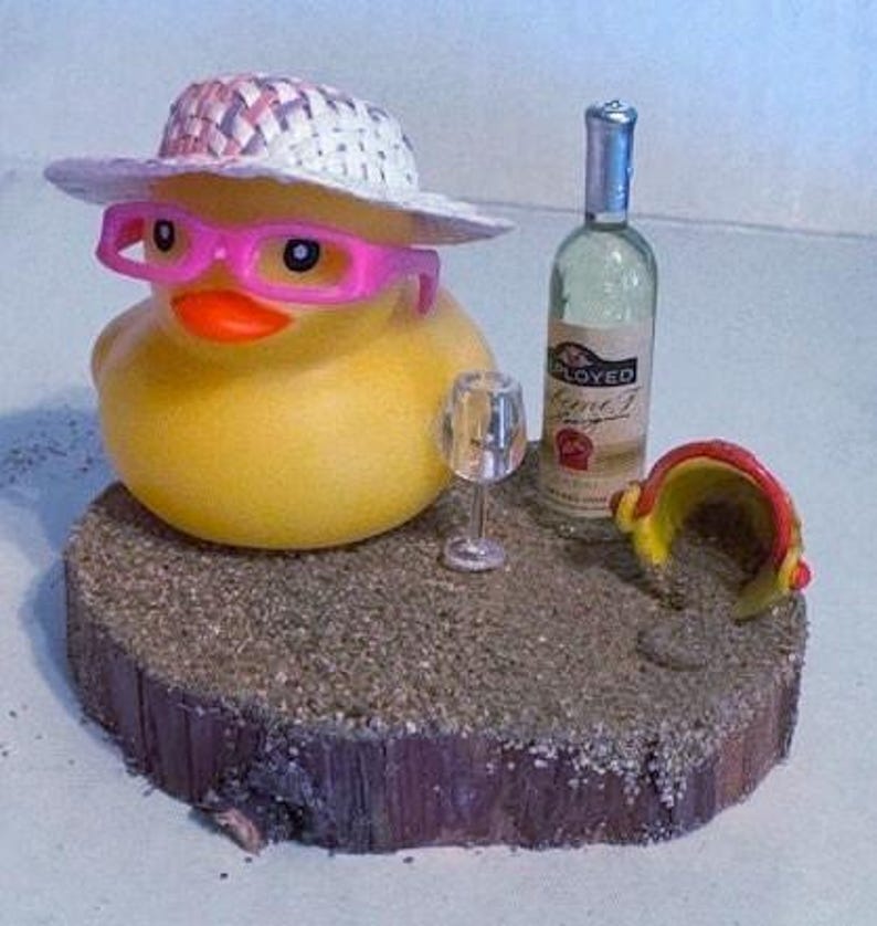 May include: A yellow rubber duck wearing a straw hat and pink glasses, next to a wine bottle and a small wine glass. The duck is on a wooden base with sand, and a small red and yellow beach bucket.