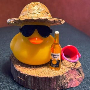 May include: A yellow rubber duck wearing sunglasses and a straw hat, holding a miniature Corona Extra beer bottle. The duck is on a wooden base with sand and a small pink bucket.