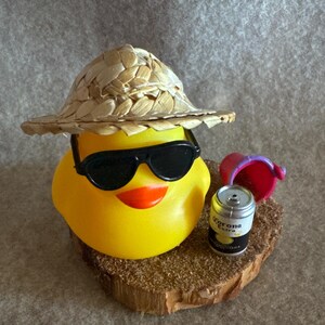 May include: A yellow rubber duck wearing black sunglasses and a straw hat. The duck has an orange beak and is sitting on a small wooden base with a miniature can of Corona Extra beer and a red bucket.