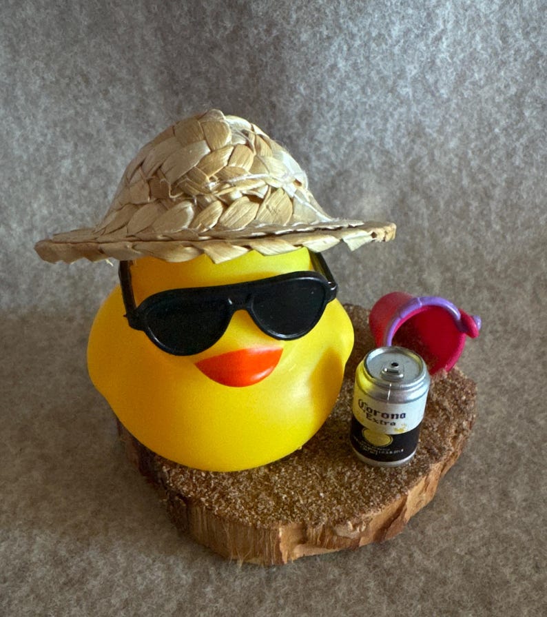 May include: A yellow rubber duck wearing black sunglasses and a straw hat. The duck has an orange beak and is sitting on a small wooden base with a miniature can of Corona Extra beer and a red bucket.