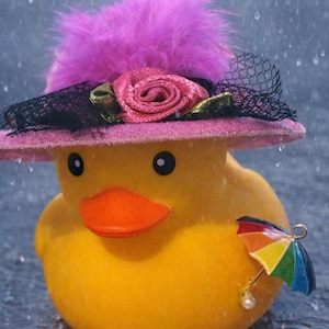 May include: A yellow rubber ducky wearing a pink hat with a rose and a black veil, and a rainbow umbrella. The hat has a pink feather and the duck has an orange beak and black eyes. The background shows raindrops.