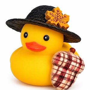 May include: A yellow rubber duck wearing a black textured hat adorned with an orange maple leaf. The duck holds a plush acorn featuring a red, white, and brown plaid pattern. The duck has black eyes and an orange beak, presented against a white background.