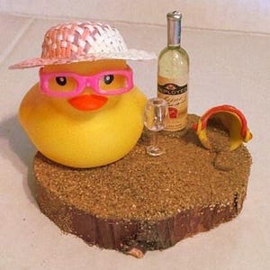 May include: A yellow rubber duck wearing pink glasses and a straw hat. A miniature wine bottle and glass are next to the duck on a sand-covered wooden base. A small yellow and red bucket is also on the base.