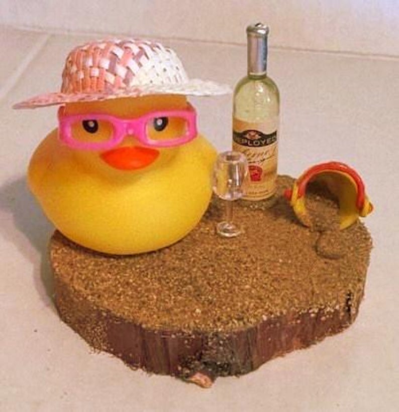 May include: A yellow rubber duck wearing pink glasses and a straw hat. A miniature wine bottle and glass are next to the duck on a sand-covered wooden base. A small yellow and red bucket is also on the base.
